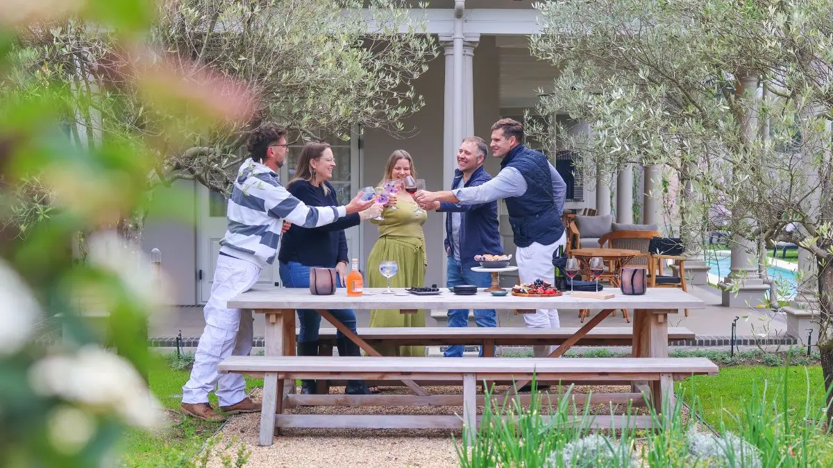 a group of people at a picnic table in a garden, making a toast